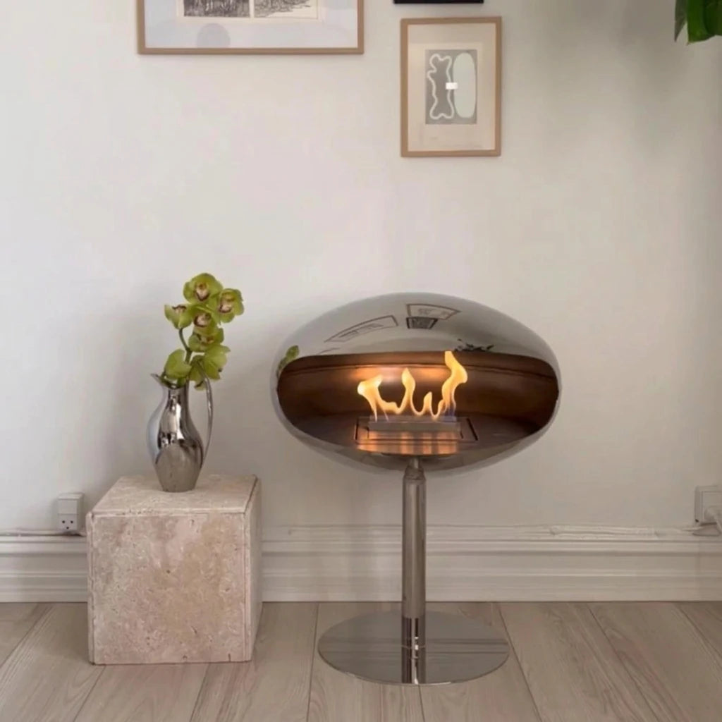 Polished steel Cocoon Pedestal bioethanol fireplace against a white wall with framed art and a silver vase with an orchid