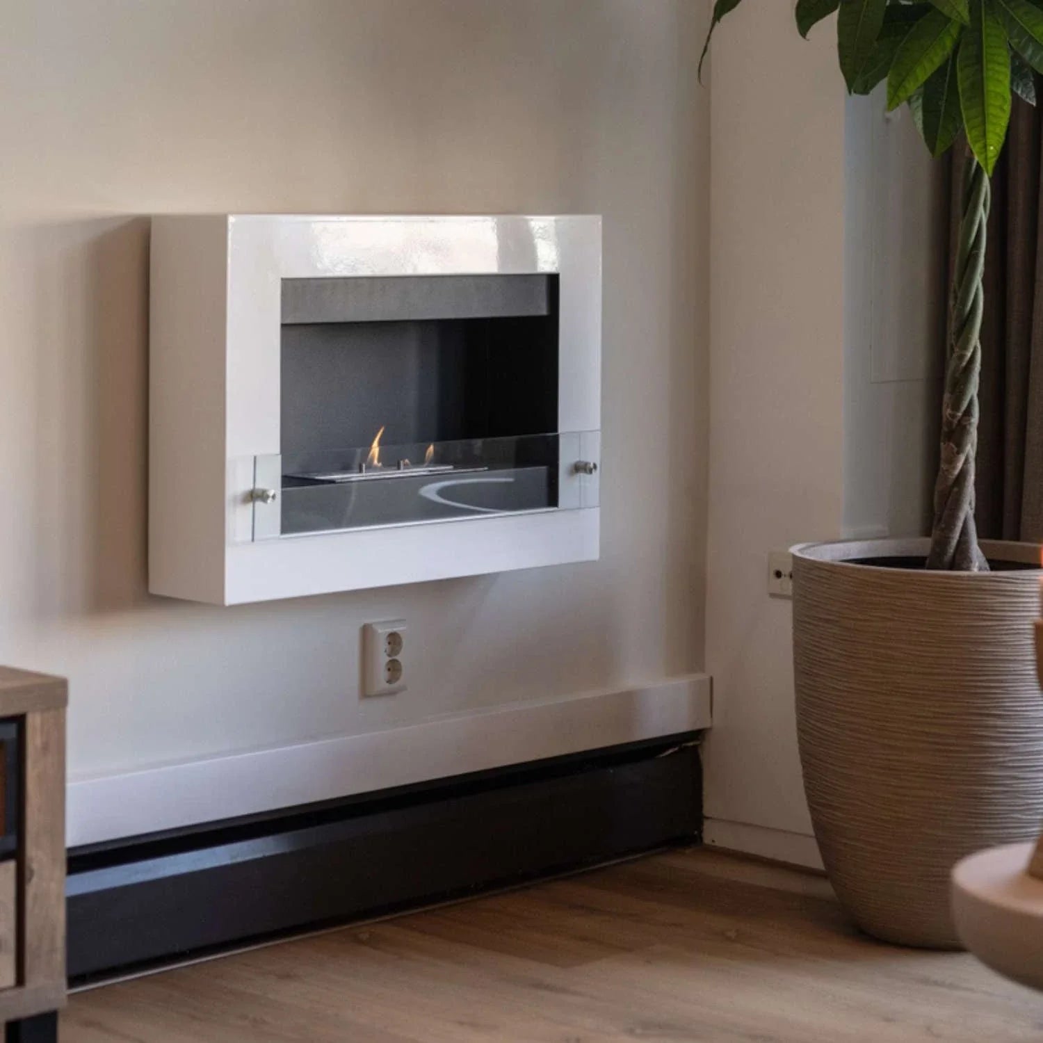 White wall-mounted bioethanol fireplace lit in a cosy living room with a tall potted plant beside it